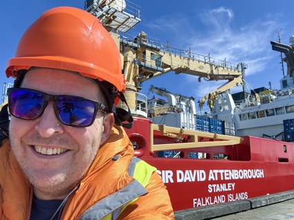 Crew member in protective clothing standing on the deck of the research vessel Sir David Attenborough, with ship equipment and icy surroundings in the background.
