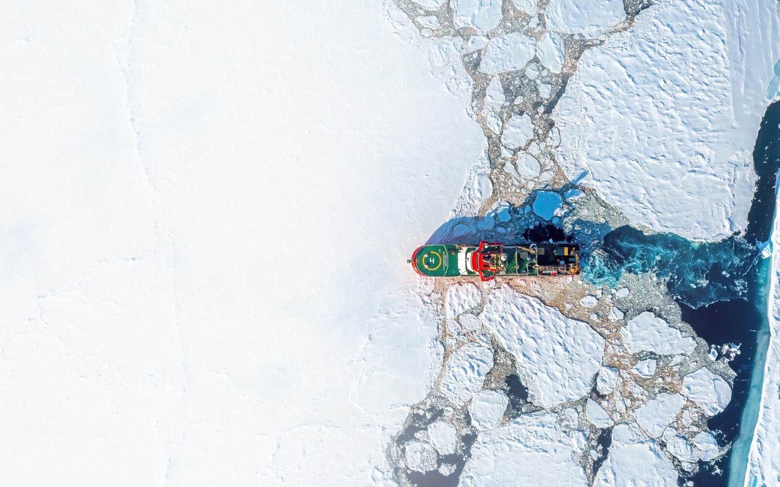 Top-down aerial view of a red ice-capable research vessel moving through cracked sea ice, with dark open water trailing behind.