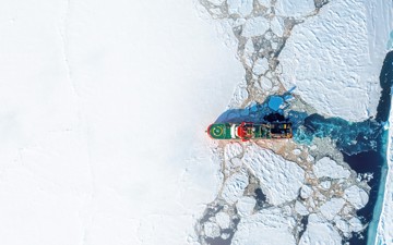 Top-down aerial view of a red ice-capable research vessel moving through cracked sea ice, with dark open water trailing behind.