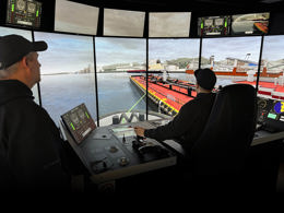 Tugboat simulator environment showing a tug assisting a large vessel in port, viewed from inside a simulated wheelhouse with screens displaying the harbour scene.