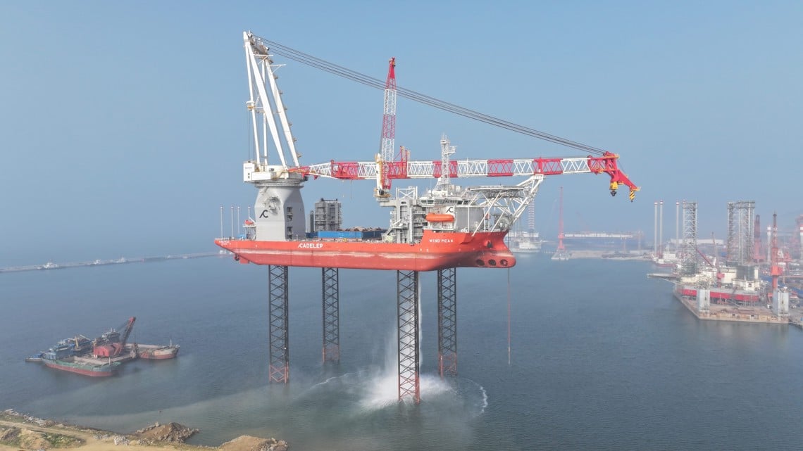 A photo shows a large wind turbine installation vessel jacked up on its legs with the hull above the sea in a commercial harbour